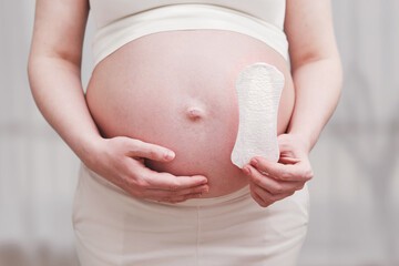 Une femme enceinte habillée d'un t-shirt blanc et d'une jupe blanche, tenant une serviette hygiénique dans une main et enveloppant son ventre de l'autre.