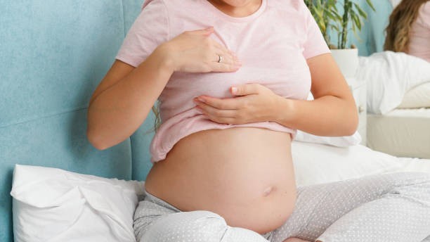 Une mère portant un t-shirt rose et un pantalon en coton, assise sur une literie blanche, en détresse à cause de la douleur à la poitrine.